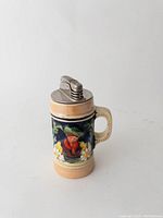 Front view of the ceramic table lighter shaped as a German stein with colorful relief decoration depicting a man in traditional clothing, flowers, and leaves. Metal lighter mechanism on top.