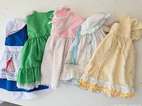 Five vintage baby dresses laid flat showing variety of styles: blue with sailboat appliqué, green, pink floral, white bonnet with blue trim, cream with yellow floral embroidery.