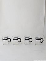 Four clear glass coffee cups with black handles and black bands around the top portion, displayed in a row on a white background.
