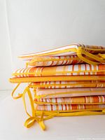 Stacked view showing eight rectangular outdoor seat cushions with orange, yellow, and white stripes and yellow ties.