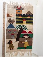 Full view of the mid century woven wall hanging showing various abstract colorful motifs including animals, houses, mountains, and figures on a beige background with fringed edges.