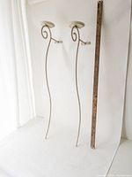 Two white swirly metal wall sconces are displayed standing upright leaning against a white backdrop. Positioned next to a yardstick for size reference, approximately 36 inches tall.