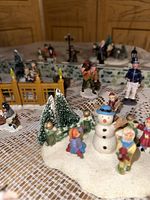 Close-up of snow-covered scene with snowman, children figurines, pine trees, and part of yellow fence gate visible
