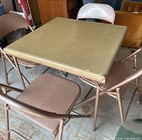 Card table with a greenish-brown laminate top and metal folding legs, surrounded by four folding chairs.