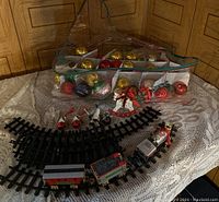 Photo showing Christmas ornaments including gold, red balls stored in compartmentalized transparent bag, Christmas train set with locomotive and cars on black plastic curved tracks, various small themed ornaments including angels and Grandma & Grandpa