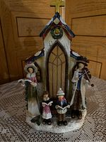 Front view of Christmas carolers figurine group standing in front of church with golden cross and wreath, captured indoors on lace tablecloth.