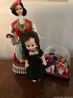 Photo showing a doll in red and white traditional attire with gold coin decorations and a smaller doll with black braided hair in front of it.