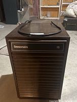 Front view of Seasonaire 15 dehumidifier showing the brand and model number on vented front panel with power cord on top, placed on concrete floor.