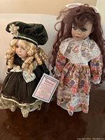Two Vanessa Ricardi collector dolls on stands, one in a black velvet dress with lace, blonde curly hair, and a black hat; the other in a floral patterned dress with lace collar and long reddish-brown hair.