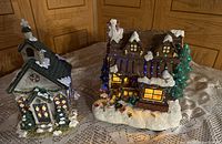 Photo showing both lighted miniature buildings, a log cabin and a chapel, on a table with wood panel background