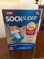 Front of the box showing the product name 'Sock Slider', large image of the device, and usage benefits including no bending or straining, and two-piece design made in USA.