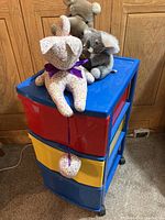 Three drawer plastic organizer with three stuffed animals on top: floral-patterned cat, gray koala bear, brown teddy bear. Organizer is blue with red and yellow drawers and black wheels.