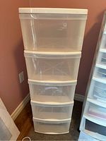 Front view of white plastic storage tower with five translucent drawers on wheels against a red wall and wood floor.