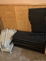 Five black plastic shelving panels stacked with black tubular legs next to them, showing the unassembled shelving unit parts