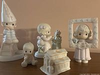 Five white porcelain Precious Moments figurines arranged on a surface, including a girl with dunce cap, baby, girl with puppy, girl in picture frame, and a church building.