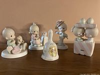 Five Precious Moments porcelain figurines arranged in a row on wooden surface including a musical bell in the center.
