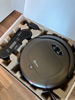 Robot vacuum and control panel in molded cardboard packaging, showing main unit shape and buttons.