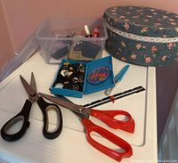 Two pairs of scissors, clear plastic sewing container, blue box of buttons, ruler, and floral sewing box visible on white surface.