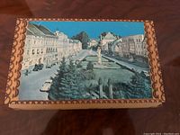Top view of music box lid showing a color photo of a European town square with buildings, cars, trees, and statues.