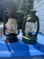 Two coal oil lanterns side by side on outdoor surface, showing overall condition and size comparison