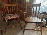 Two solid wood chairs positioned in a room, showing turned spindle legs and armrests, one chair without cushion and one with a patterned cushion.