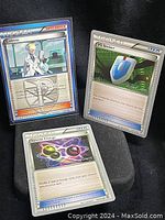 Three rare Pokémon Trainer cards shown vertically stacked with visible character art and blue and silver borders, in protective sleeves on a black surface.