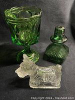 Green glass goblet, green perfume/oil bottle, and clear glass Scottish Terrier figurine together on black background.