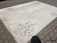 Full view of light cream wool carpet laid on a brick patio, displaying floral embroidery and scroll embossed patterns with fringe on one edge.