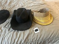 Two summer hats laid flat on a bed: a black woven wide-brim hat with frayed edges and a yellow and beige wide-brim sun hat with a clear inner band.