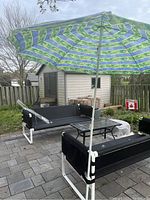 Full view of the patio furniture set with the green and blue striped beach umbrella open, showing the couch, chair, and glass-top table on a paved patio.