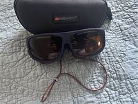 Polarized sunglasses resting on quilted surface, black cord attached, case visible behind glasses