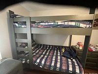 Grey bunk bed frame with a built-in ladder, showing front and side views with mattresses and plaid bedding.