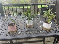 Three gold metal hanging plant holders on outdoor metal table, each holding a small plant pot. Showing set of three with geometric triangular frames and round bases.