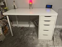 Front and side view of the white IKEA desk showing five drawers on the right side and a round cylindrical metal leg on the left side. Desk surface is clean and holds a lava lamp, figurine, and clock.