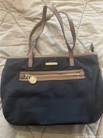 Front view of black Michael Kors tote bag with tan leather handles and trim showing front zip pocket and logo plaque.