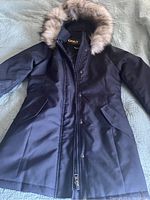 Front view of dark winter coat with fur trimmed hood laid out flat to show details such as pocket placement and closure style.