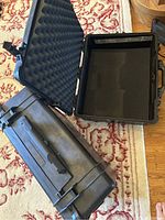 Two black Pelican cases placed on a patterned rug, one case open showing egg crate foam on lid and moveable foam interior in bottom section.