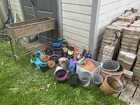 Wicker planter stand, assorted pots, metal baskets, and containers outside on grass next to house, with stacked bricks in background