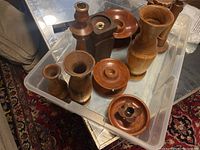 Seven turned wood items arranged in a clear plastic bin, visible wood grain and different shapes including candle holders, vases, and a box.