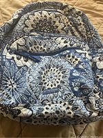Front view of Vera Bradley quilted backpack with blue and white floral pattern, showing front zippered pocket and fabric tag