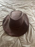 Brown vintage leather cowboy hat displayed on a beige quilted surface, showing the top and brim with visible wear and creases.