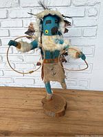 Full view of the Kachina statue standing on wooden base, showing blue painted face mask, fur clothing, suede skirt, and feathers on back.