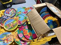 Box filled with colorful lightweight plates featuring tropical and character designs piled in a cardboard box on a yellow plastic bin.