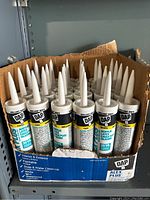 Full front view of the case containing multiple white tubes of DAP Alyx Plus Acrylic Latex Caulk with pointed nozzles.