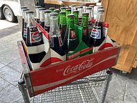 Wooden Coca-Cola crate filled with an assortment of empty glass soda bottles including Pepsi and 7 Up, showing front and side of crate and bottles.