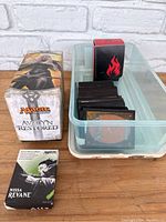 Photo showing Avalon Restored Magic The Gathering box with fantasy character art and transparent container filled with loose Magic The Gathering cards. A smaller black box with red flame icon is visible inside the container. Front portion of the container shows card backs with classic Magic card back design.
