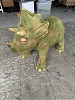 Photo showing full side and front view of green dinosaur ride-on toy with fabric exterior and beige claws, housed in a garage-like setting.