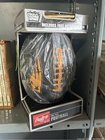 Full view of the packaged Rawlings full size black football featuring New England Patriots star logo and laces in orange, inside original box packaging.