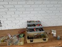 Photo of open tacklebox filled with assorted fishing lures and fishing accessories laid out on wooden surface with brick wall background.