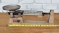 Full view of the triple beam balance scale showing the metal weighing pan, aluminum beam with three sliding weights, and the beige base with measurement scale alongside a measuring tape.